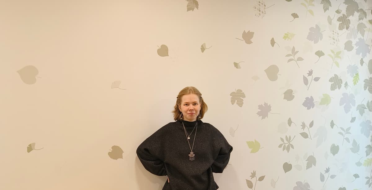 A person in a black top is standing in front of a light-colored wall decorated with leaf patterns. They are facing the camera with their hands behind their back.