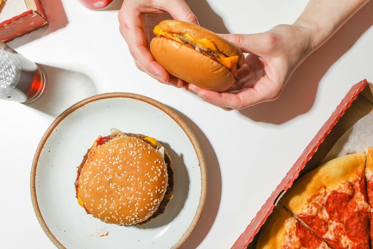 en hamburgare syns i en persons händer, på en tallrik finns en annan hamburgare och i en kartong finns pizza