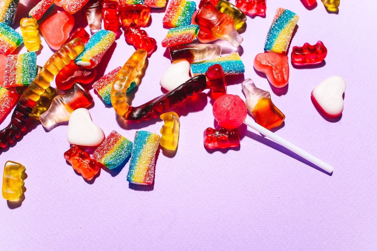 Colorful gummy candies, sour rainbow strips, heart-shaped sweets, and a lollipop are scattered across a light purple surface.