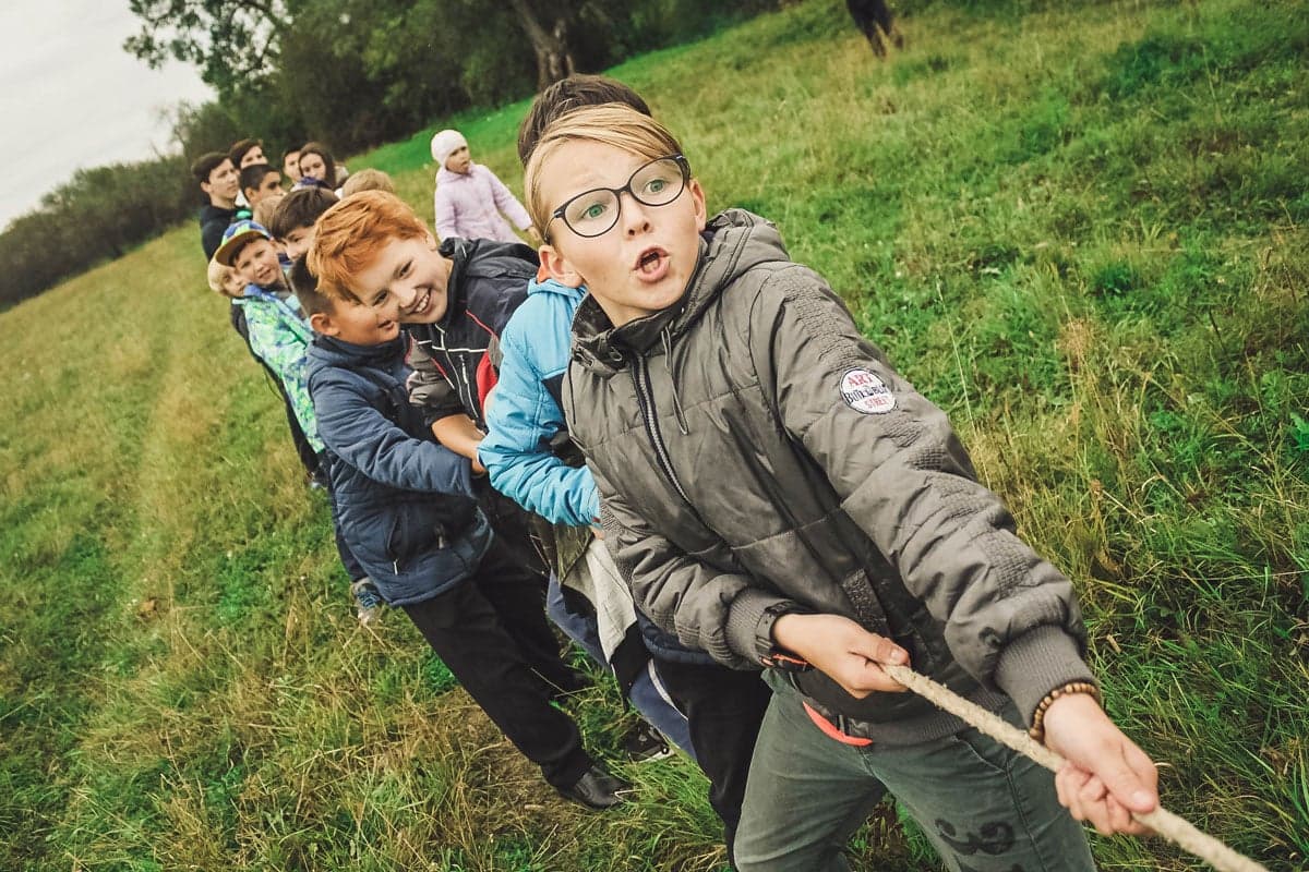 En grupp barn deltar i dragkamp på en gräsbevuxen äng, med entusiastiska ansikten och samarbete som fokus.