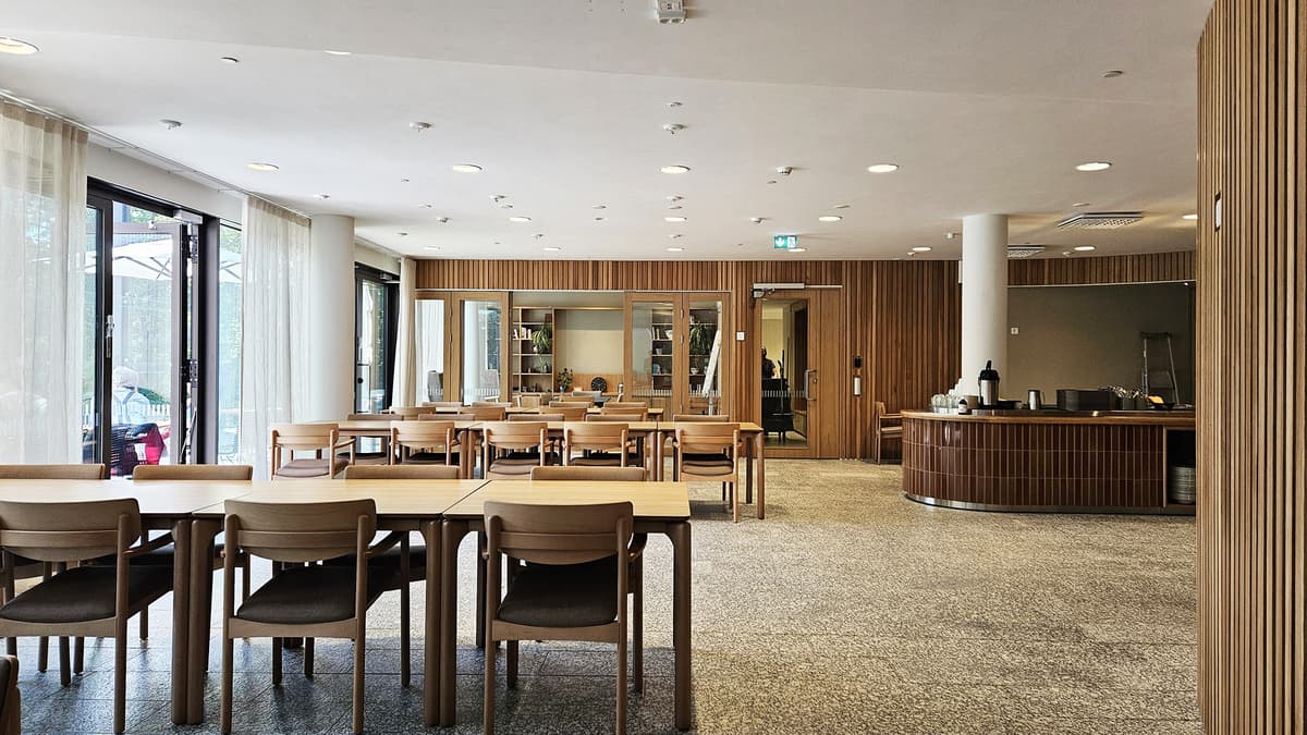 Ljus matsal med träbord och stolar, stora fönster, träpanel på väggarna och en serveringsdisk.