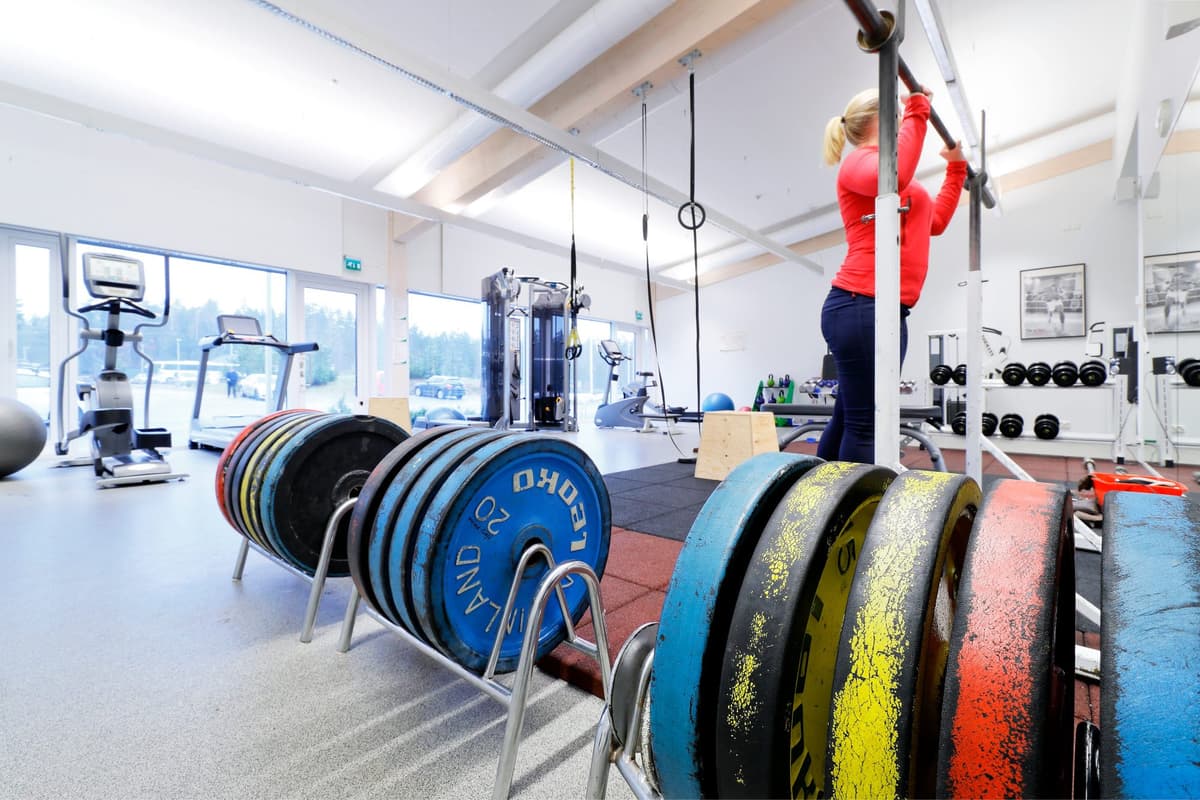 En person tränar på en skivstång i ett välutrustat gym med fria vikter, träningsmaskiner och konditionsutrustning. Ljusa fönster ger en öppen och inspirerande träningsmiljö.