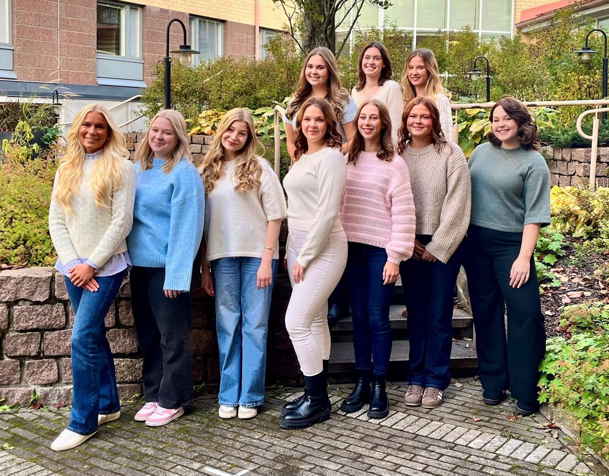 Gruppfoto av tio unga kvinnor som står i två rader utomhus framför en låg stenmur och höstgrönska. De bär stickade tröjor i ljusa och dova färger.