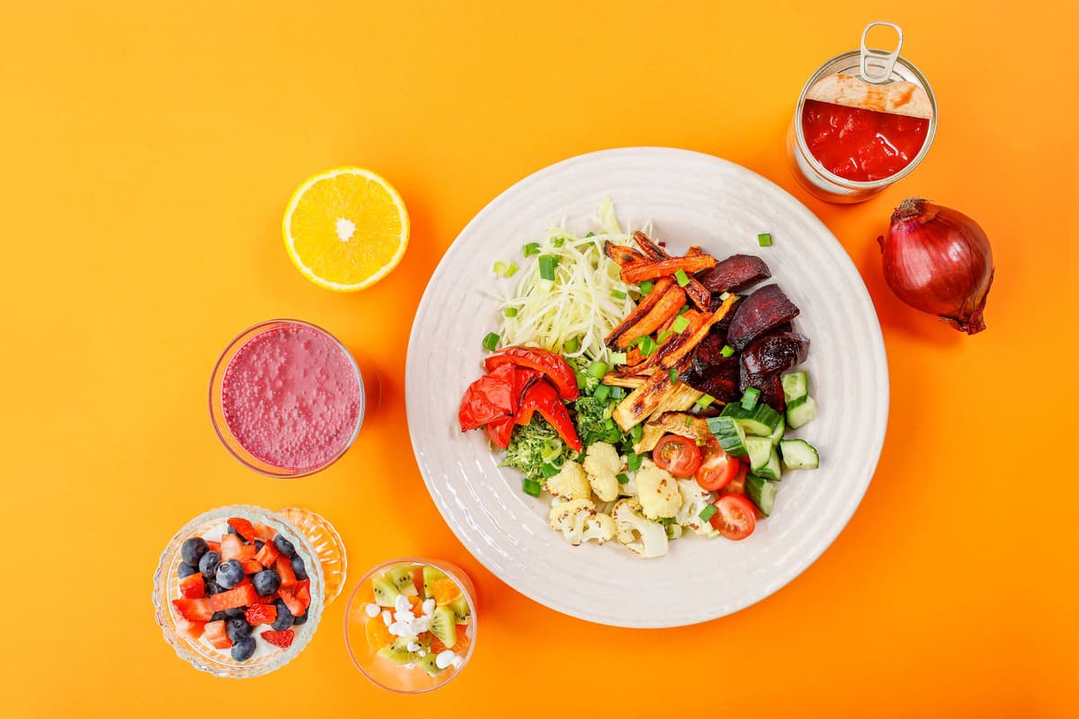 Tallrik med ugnsrostade rotfrukter, färska grönsaker och kål, omgiven av en halv apelsin, rödlök, tomatkross, bär, smoothie och fruktdessert.