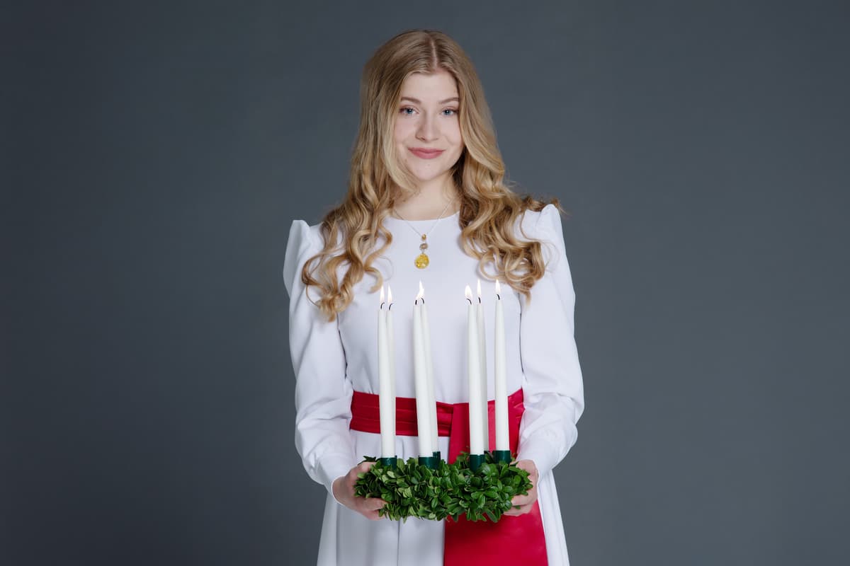Finlands Lucia, klädd i vit skrud med rött band, håller leende fram en luciakrona med brinnande ljus i.