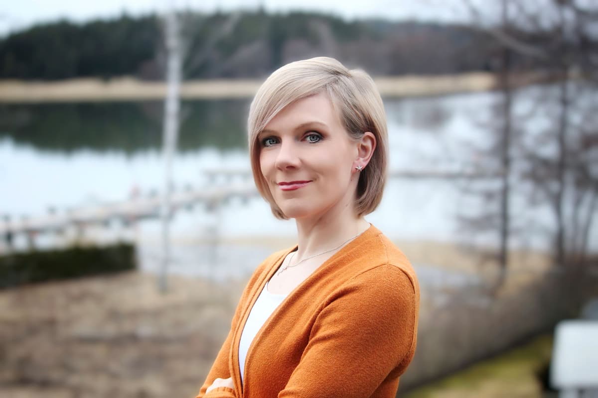 Kvinna i orange tröja står i naturen med armarna i kors och tittar rakt in i kameralinsen.