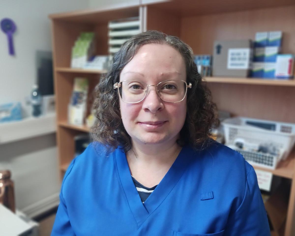 En person med glasögon och blå arbetsrock sitter inomhus framför en hylla med olika föremål. Bilden är tagen på nära håll och visar personen centrerad i motivet.