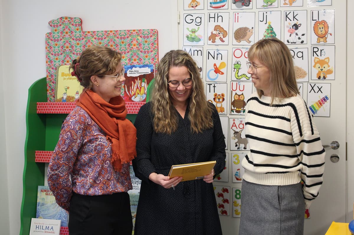 Tre kvinnor står brevid varandra och ser glatt på en barnbok.