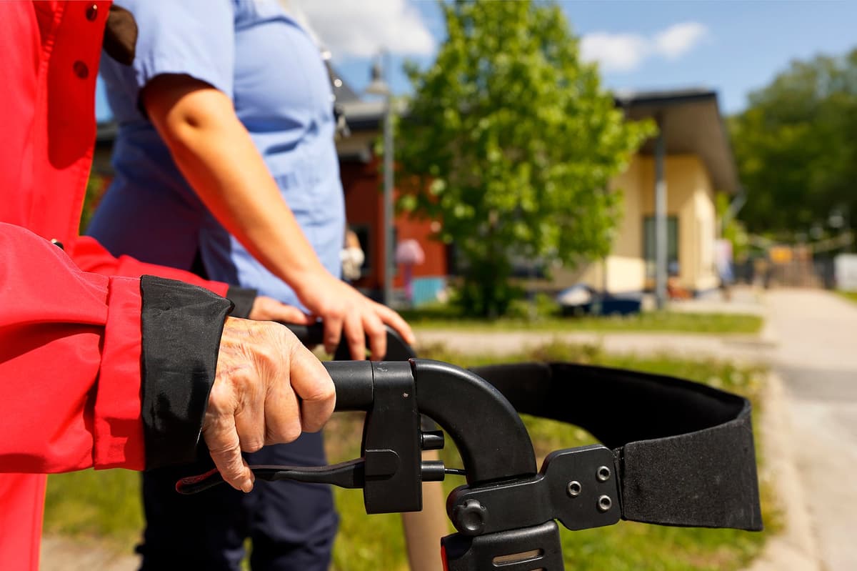 En vårdare promenerar ute i soligt väder med en äldre, som stöder sig på en rollator. 