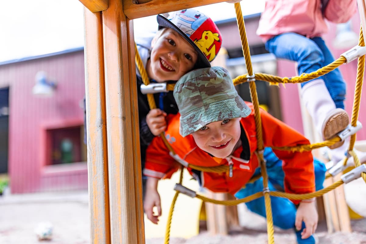 Två glada barn har klättrat upp i en klätterställning och ser glatt ner genom den. 