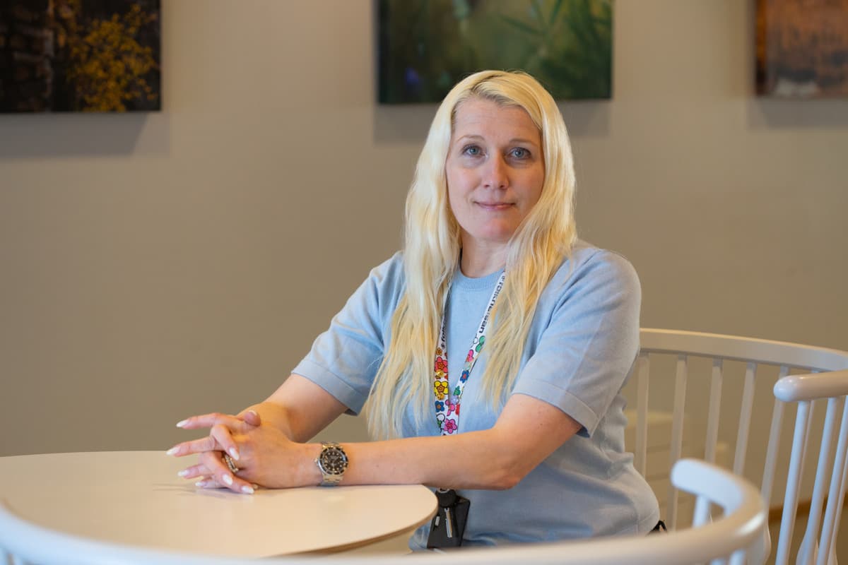 En blond kvinna sitter vid ett bord inomhus och tittar mot kameran. Hon har på sig en ljusblå tröja och ett färgglatt nyckelband, med tavlor på väggen bakom sig.