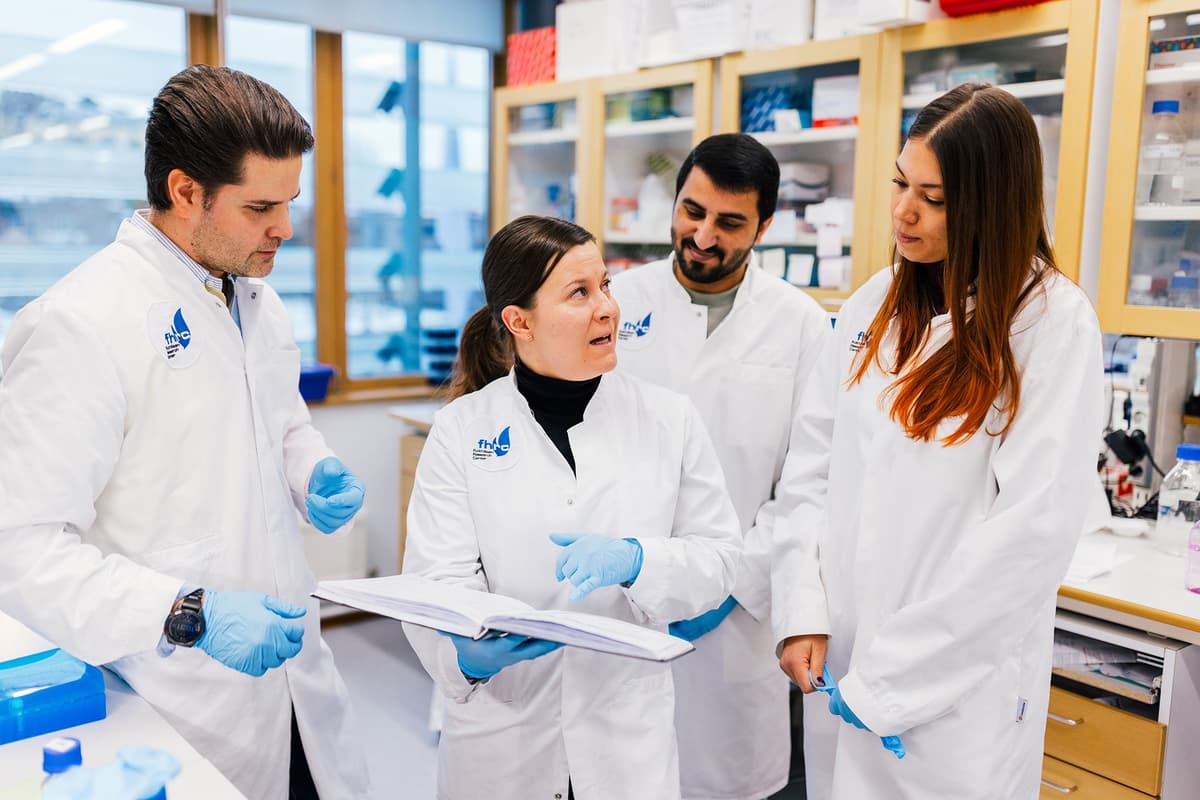 Four researchers in white lab coats and gloves stand in a laboratory, discussing an open research book. The bright and modern work environment is filled with lab equipment and large windows letting in natural light.