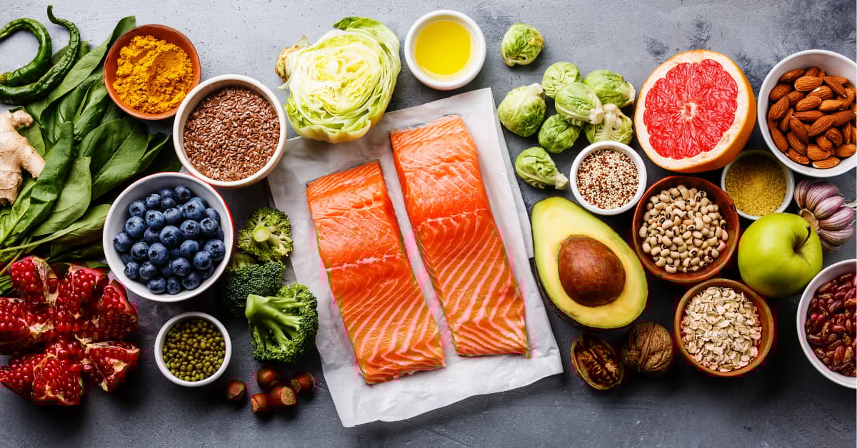 Ett urval av hälsosamma livsmedel arrangerade på ett bord. Bilden visar laxfiléer, grönsaker som broccoli, brysselkål, avokado, kål och spenat. Dessutom finns frukter som blåbär, granatäpple, grapefrukt och äpple, samt nötter, frön, bönor, havre och kryddor som gurkmeja. En skål med olivolja syns också.