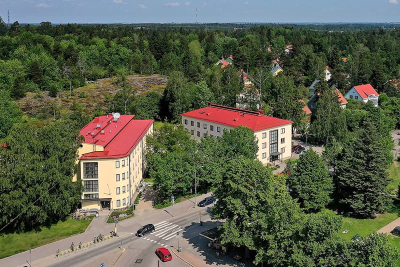 Flygbild över Folkhälsans byggnader i Kottby, omgivna av grönska och småhus. De ljusgula husen med röda tak ligger vid en vägkorsning med övergångsställe och cykelparkering. I bakgrunden syns skog och ett bostadsområde med villor bland träden.
