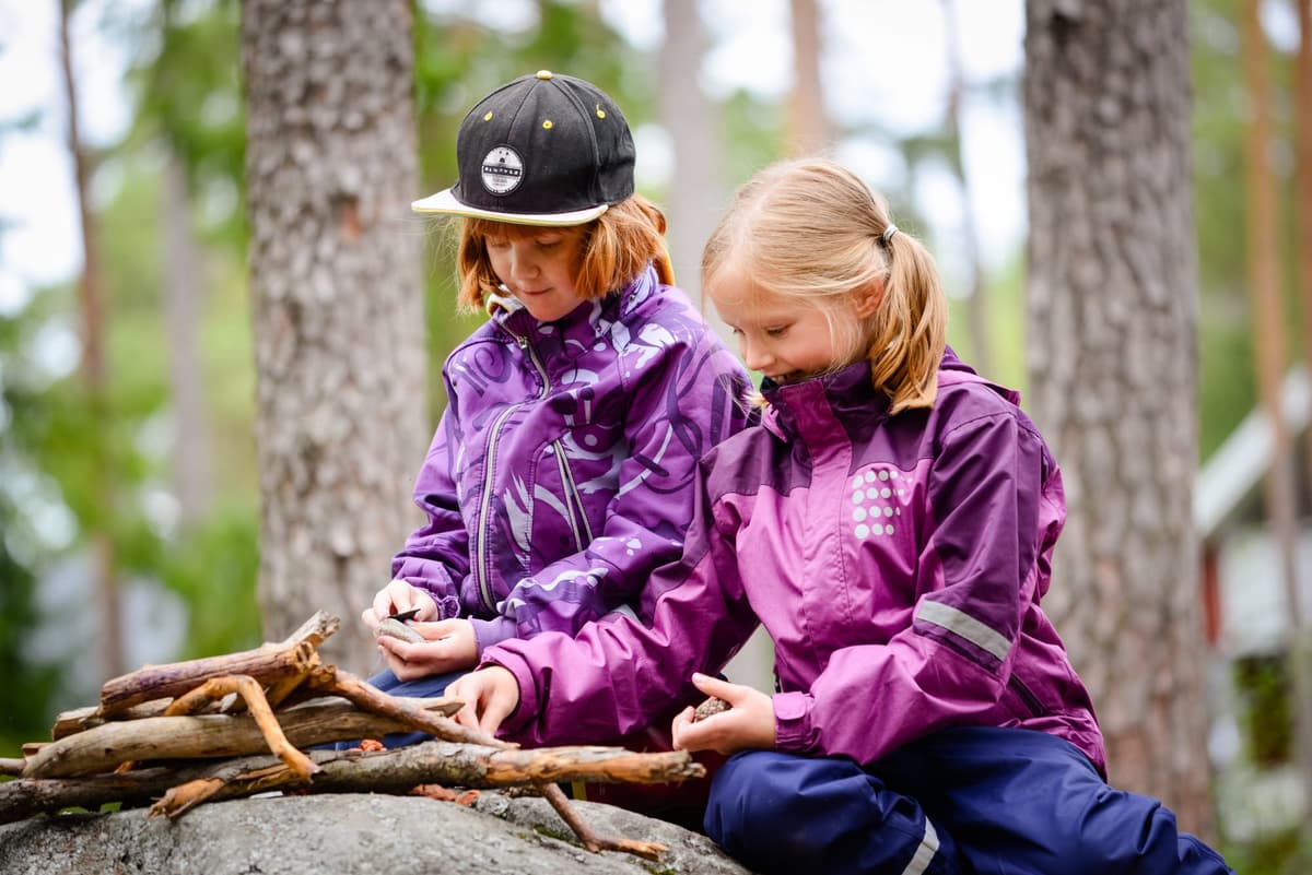 Två barn klädda i lila ytterkläder sitter utomhus och bygger med pinnar, omgivna av träd i en skogsmiljö.