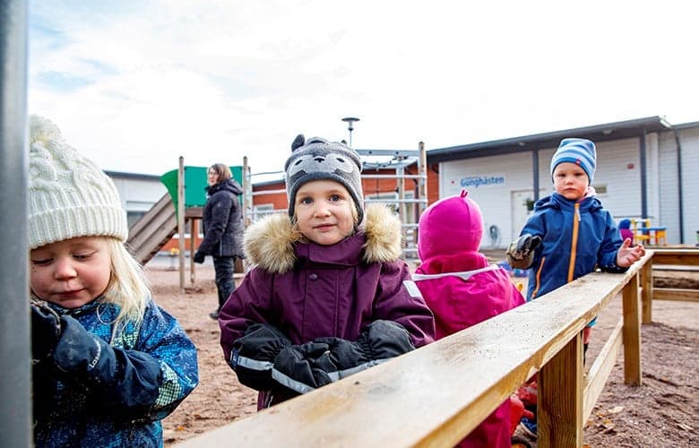 Barn som leker utomhus på en lekplats i vinterkläder. En pedagog syns i bakgrunden vid klätterställningen.