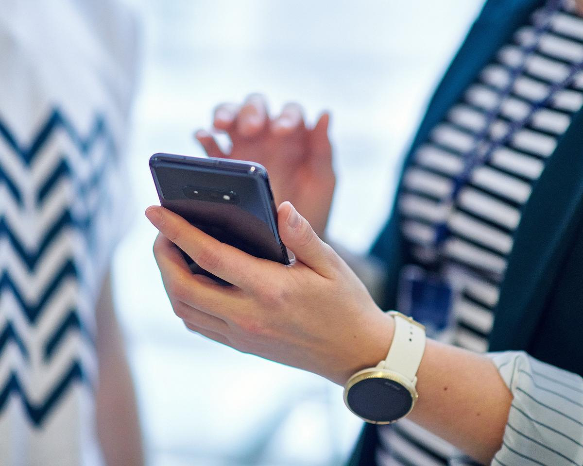A person holds a smartphone in one hand while gesturing with the other, wearing a wristwatch and a striped shirt. The background is blurred, suggesting a conversation or workplace setting.