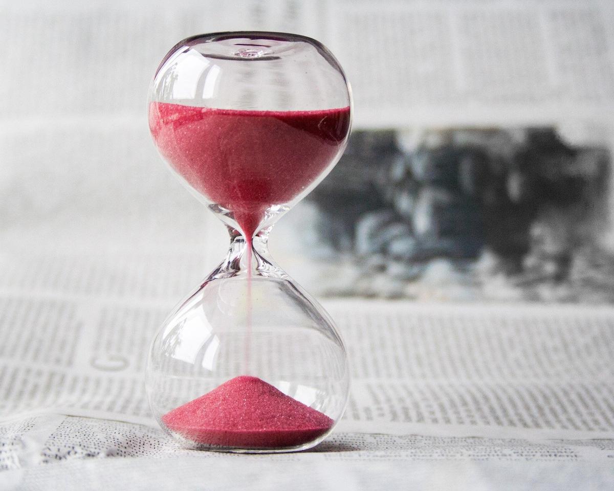 A glass hourglass with red sand sits on a newspaper as the sand flows from the top chamber to the bottom. The background is softly blurred, emphasizing the passage of time.