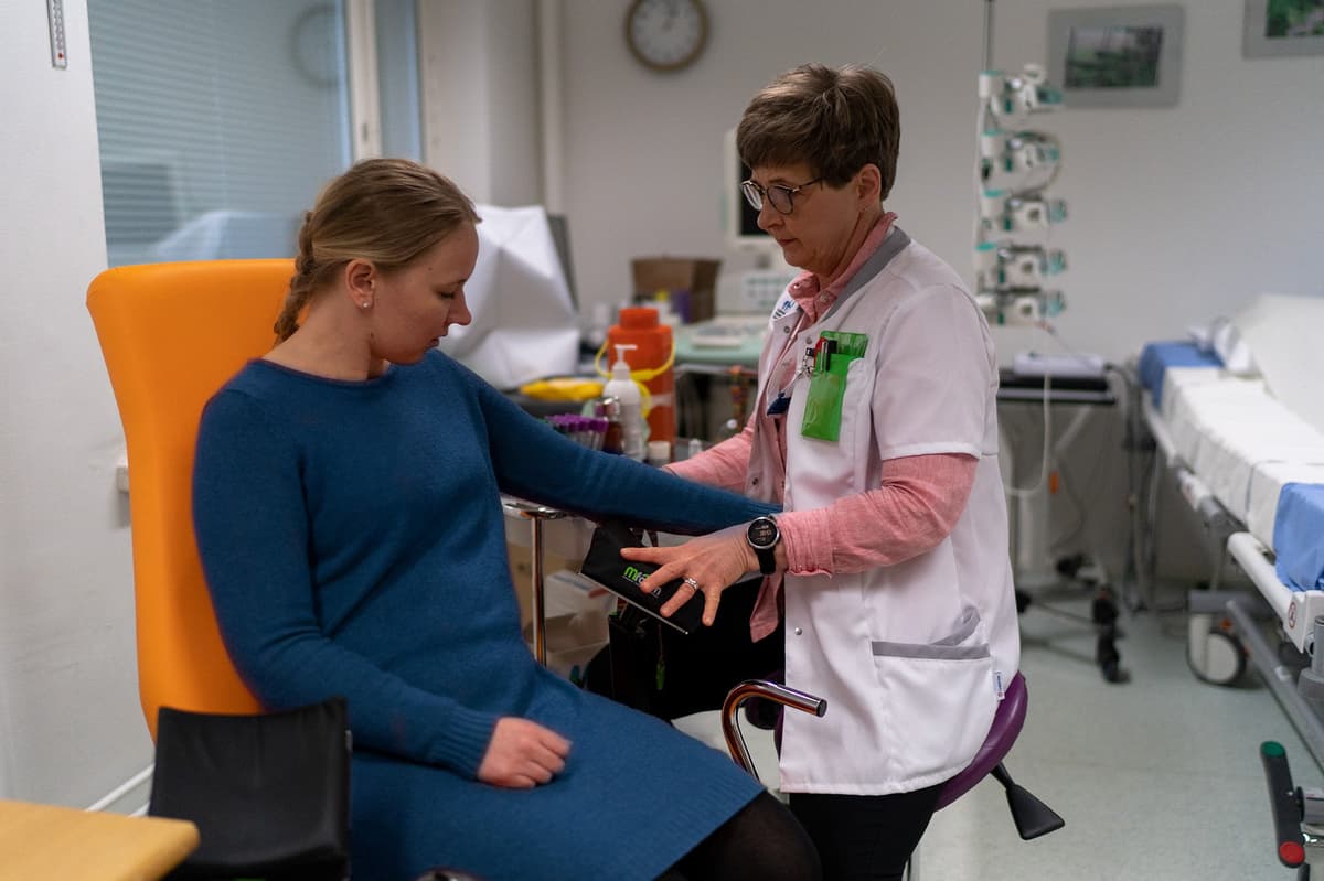 A healthcare professional in a white coat measures a woman's blood pressure as she sits in an orange examination chair. Medical equipment and supplies are visible on a nearby table and in the background, indicating a clinical or research environment.