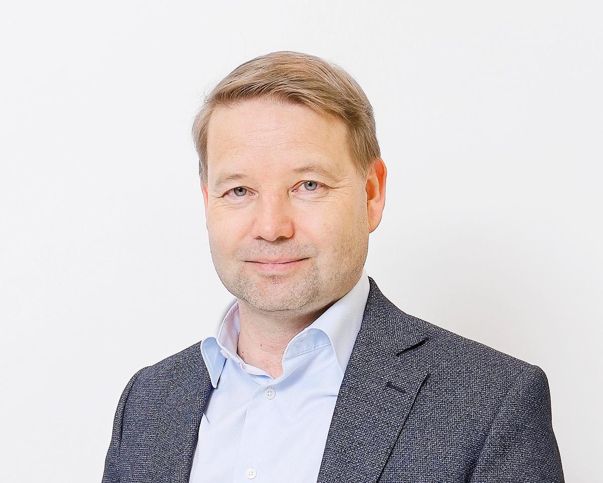 En man med kort hår står framför en vit bakgrund. Han bär en ljus skjorta och en mörk kavaj.
