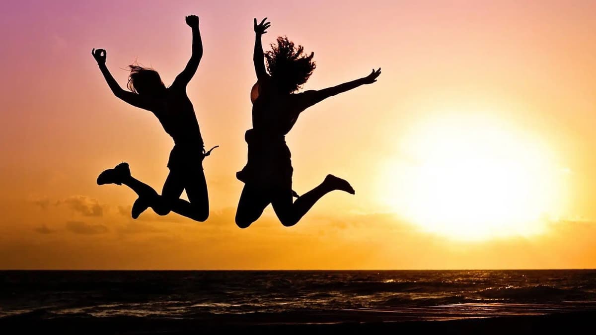 Two people are captured mid-air as they joyfully jump against the backdrop of a vibrant sunset by the sea. Their silhouettes are sharply outlined by the glowing sky, creating a lively and energetic scene.