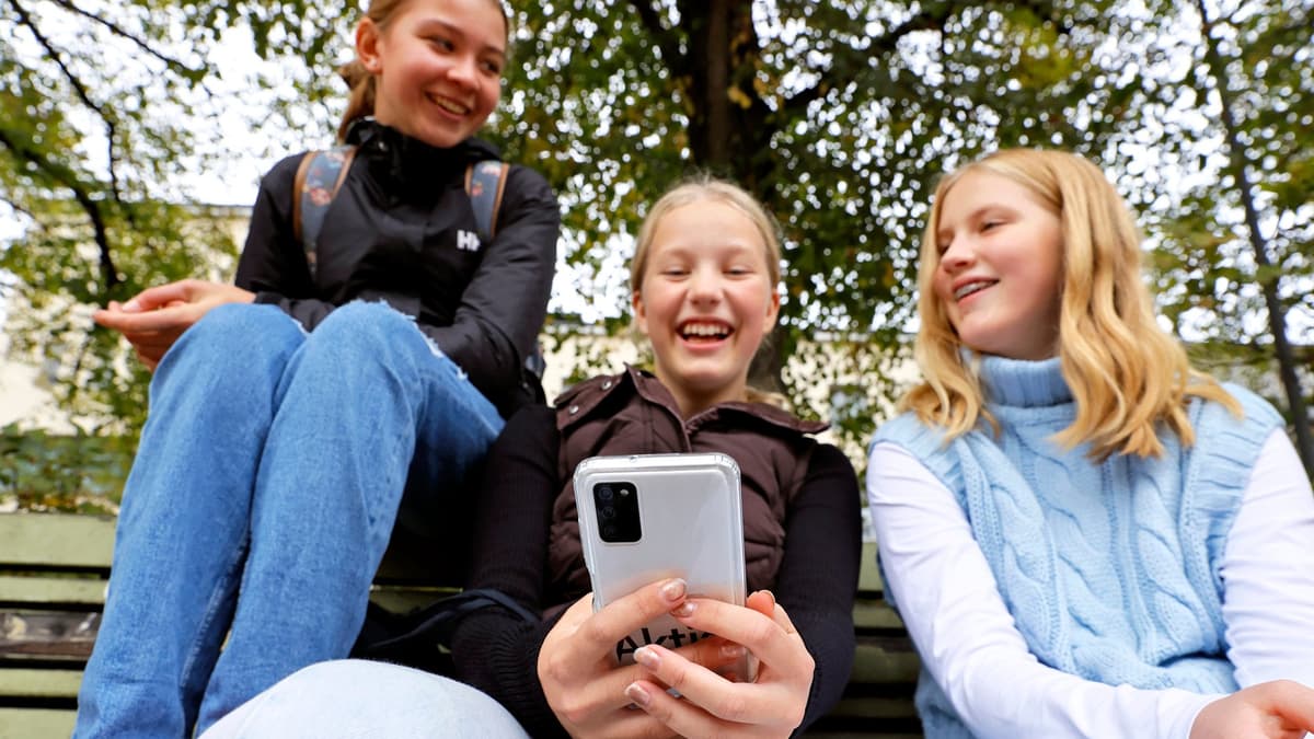 Three children sit on a bench outdoors, looking at a smartphone held by the child in the middle. Trees with leaves are visible in the background above them.