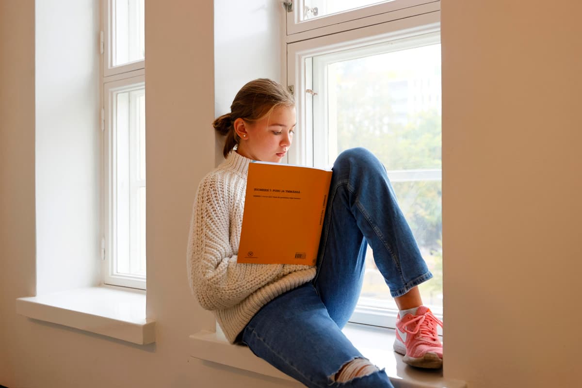 En ung person sitter i en fönsternisch och läser en orange bok. Solljuset strömmar in genom de stora fönstren och skapar en lugn och ljus stämning.