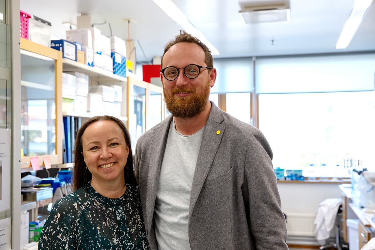 Two people stand close together in a laboratory, smiling at the camera. Lab shelves, supplies, and workbenches are visible in the bright background.