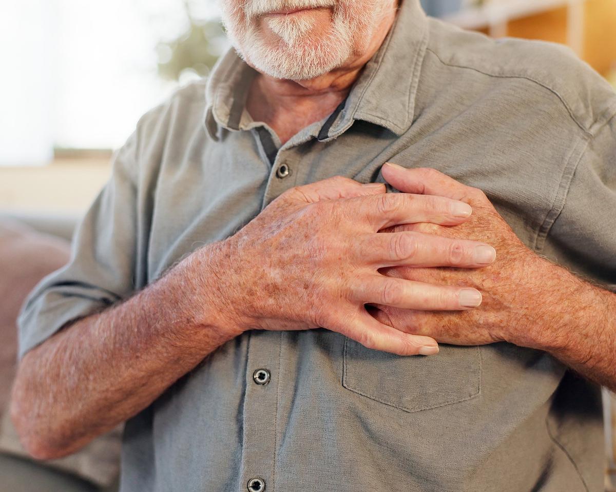 An older person in a grey shirt holds both hands against their chest. The image shows a close-up of the upper body with the face partially out of frame.