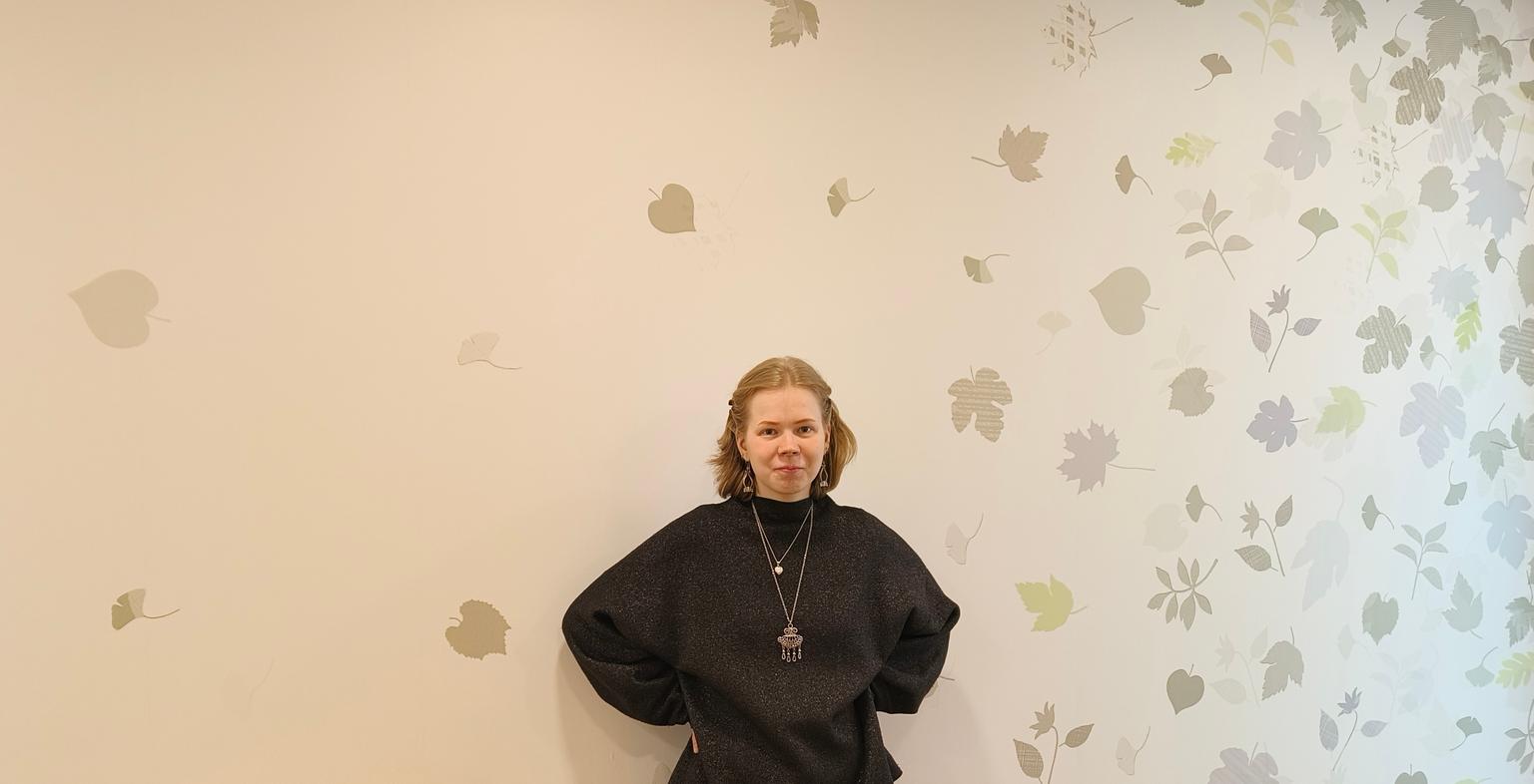 A person in a black top is standing in front of a light-colored wall decorated with leaf patterns. They are facing the camera with their hands behind their back.