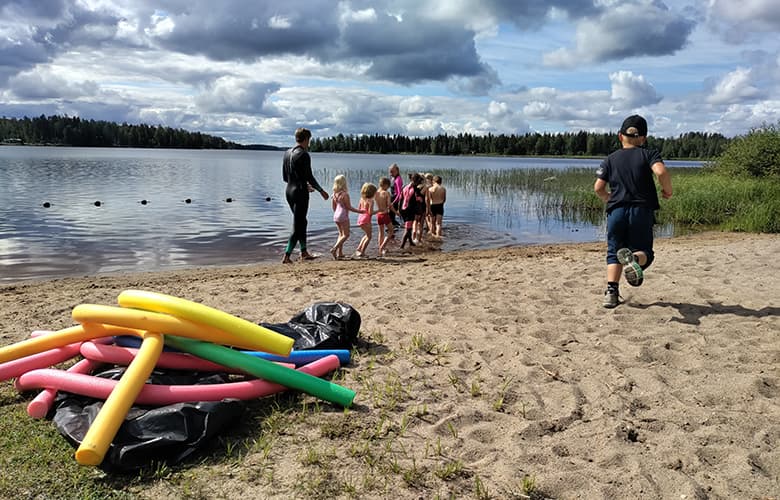 En grupp barn går ner i vattnet vid en strand, ledda av en vuxen i våtdräkt. En annan person springer mot dem från sanden. I förgrunden ligger simdynor i olika former och en sopsäck på marken. Himlen är fylld med moln, och i bakgrunden syns en sjö omgiven av skog.
