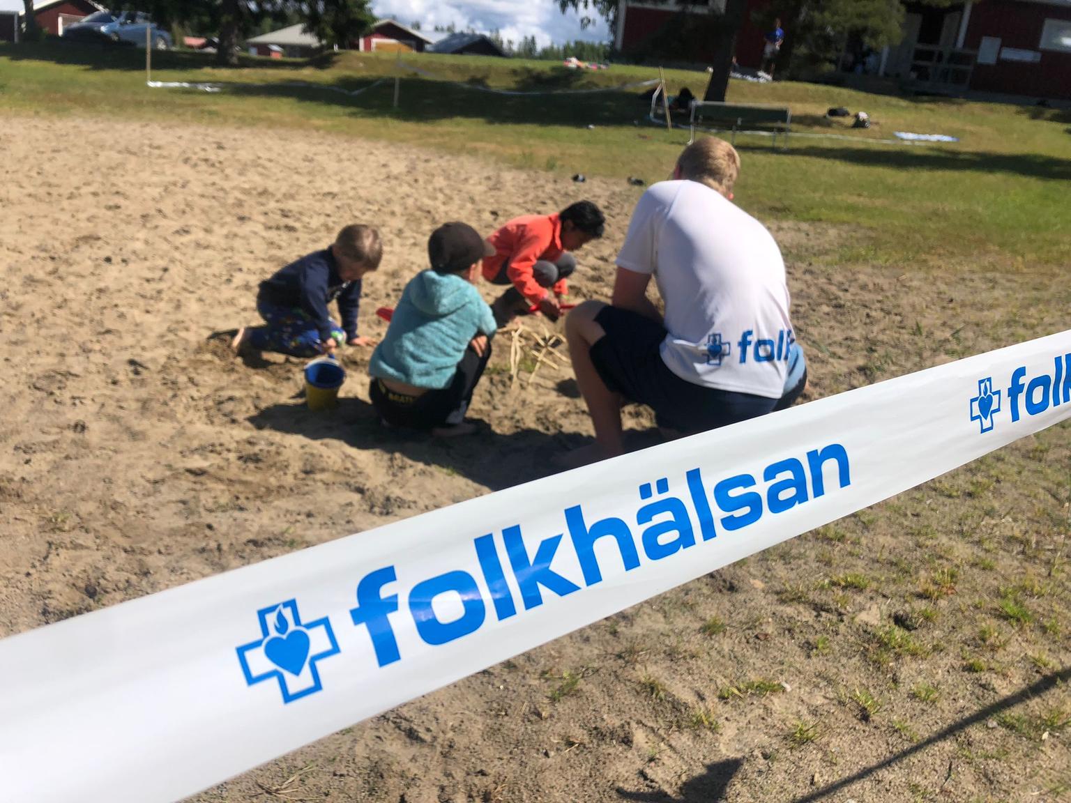 En grupp barn och en vuxen leker tillsammans i sanden, omgivna av ett avspärrningsband med Folkhälsans logotyp och namn. I bakgrunden syns gräs, byggnader och några personer längre bort.