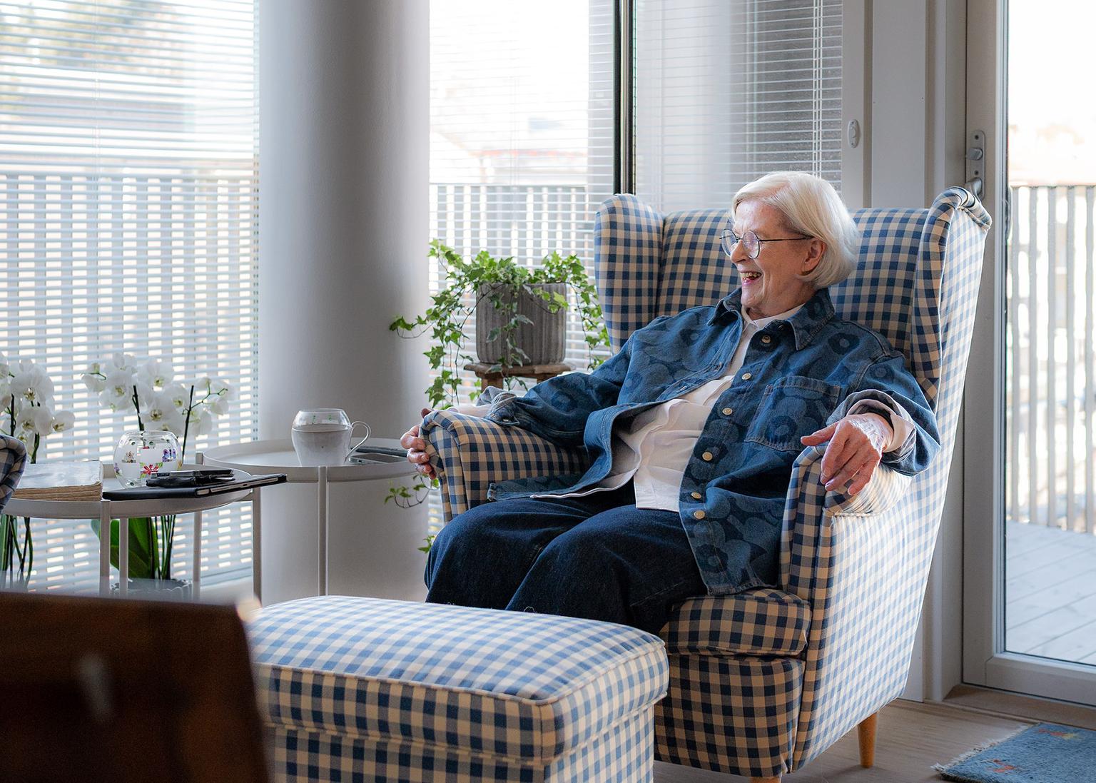 En äldre kvinna med vitt hår och glasögon sitter leende i en blårutig fåtölj vid ett stort fönster.
