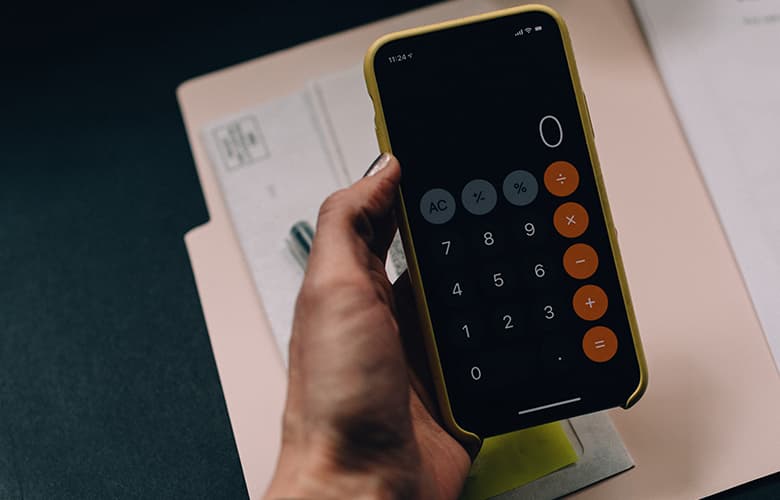 En hand håller i en smartphone med kalkylatorn öppen på skärmen, och i bakgrunden syns en skrivbordsunderlägg och dokument.