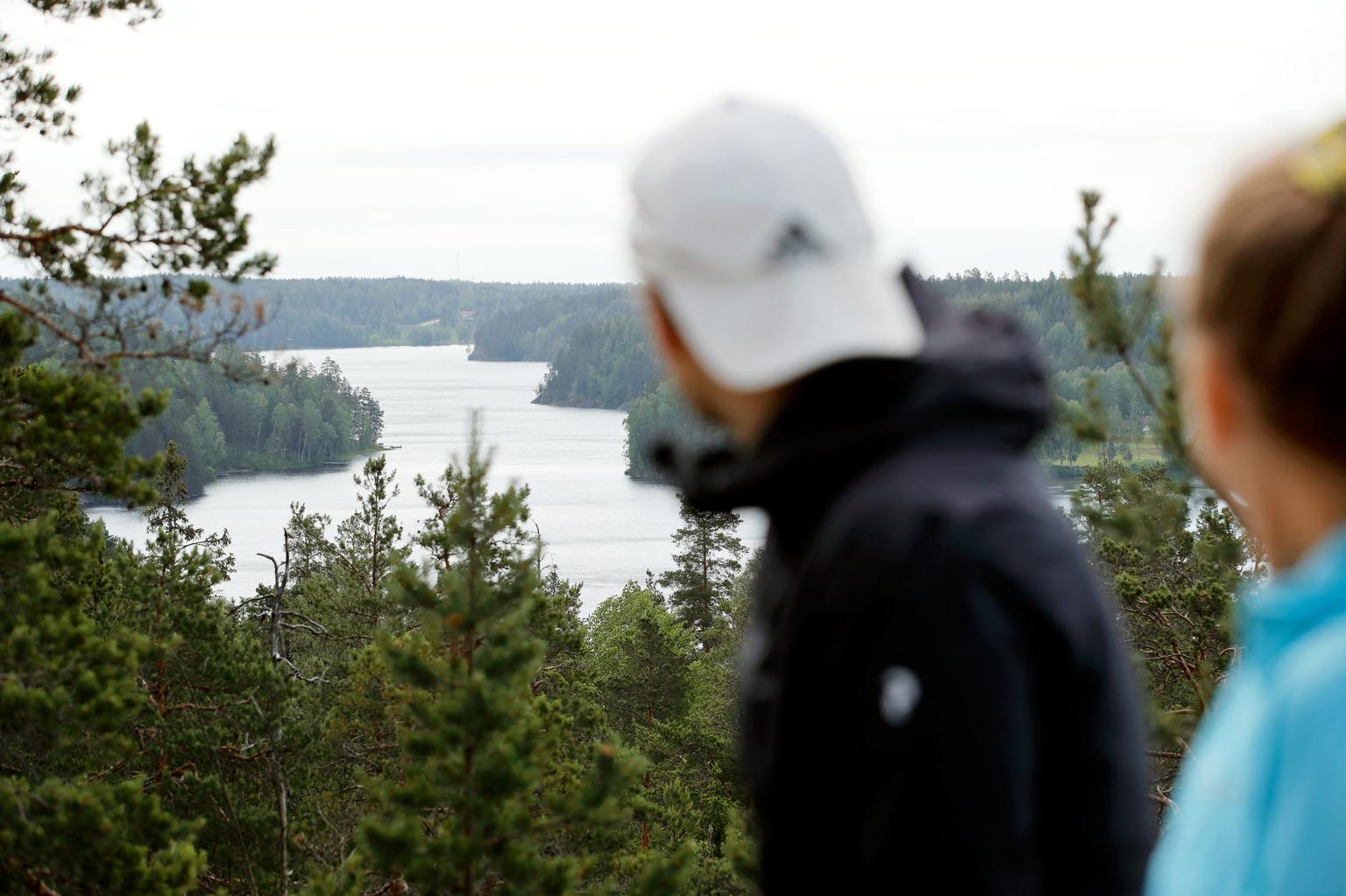 Kaksi henkilöä seisoo korkealla paikalla ja katselee kaunista järvimaisemaa, jota ympäröi metsä. Näkymä on rauhallinen ja luonnonkaunis.