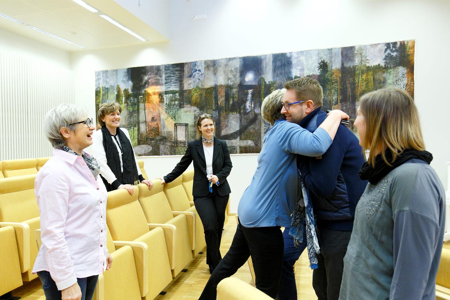 En grupp människor samlas i en ljus konferenssal med gula stolar. De hälsar varmt på varandra, skrattar och kramas. I bakgrunden syns en stor konstnärlig målning med naturmotiv. Stämningen är glad och inkluderande.