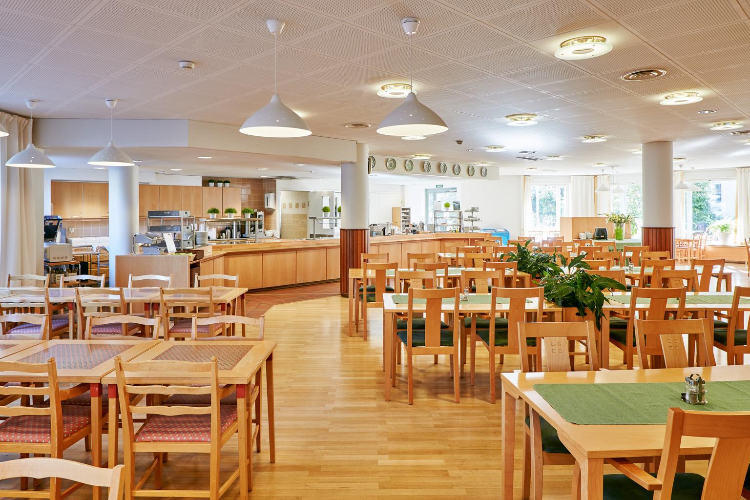 En rymlig lunchrestaurang med trämöbler och gröna bordstabletter. Restaurangen har ett ljust trägolv och stora fönster som släpper in naturligt ljus. Taklampor i vitt hänger över borden, och serveringsområdet i bakgrunden har träinredning med gröna växter som dekoration.