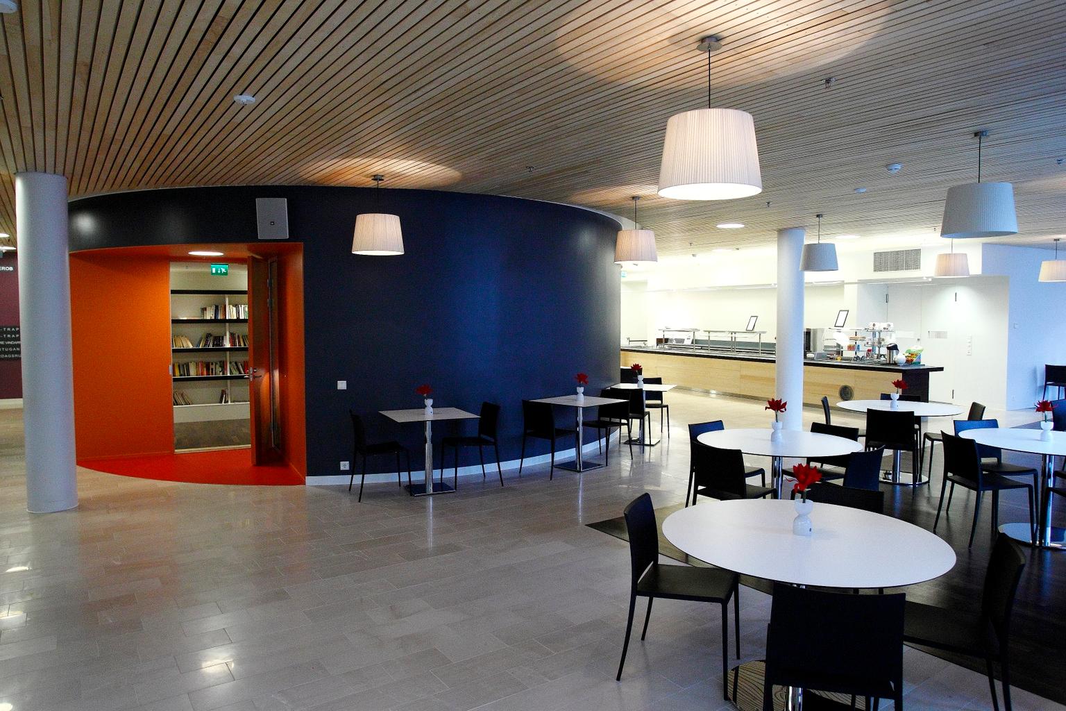En modern restaurang med träpanel i taket och vita väggar. Runda och fyrkantiga bord är omgivna av svarta stolar, och varje bord har en vit vas med röda blommor. Till vänster syns en öppning till ett bibliotek med orange dörrkarm. I bakgrunden finns en serveringsdisk.