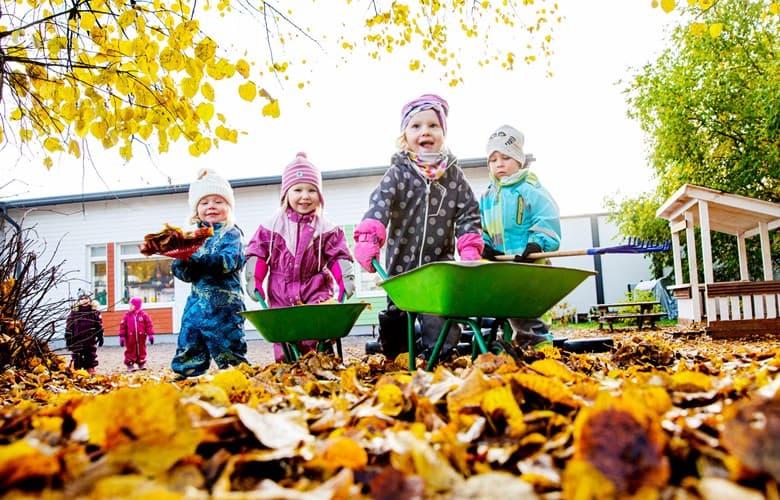 Barn leker och samlar höstlöv i skottkärror utomhus på gården.