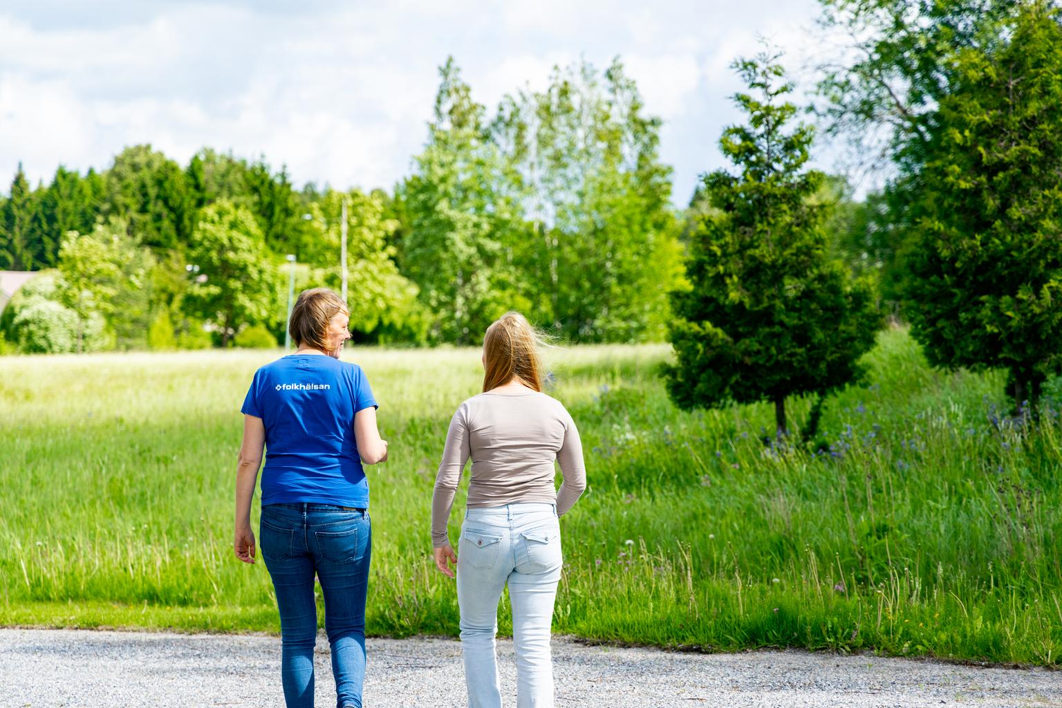 Två personer promenerar tillsammans på en grusväg i en grön naturmiljö en solig dag.
