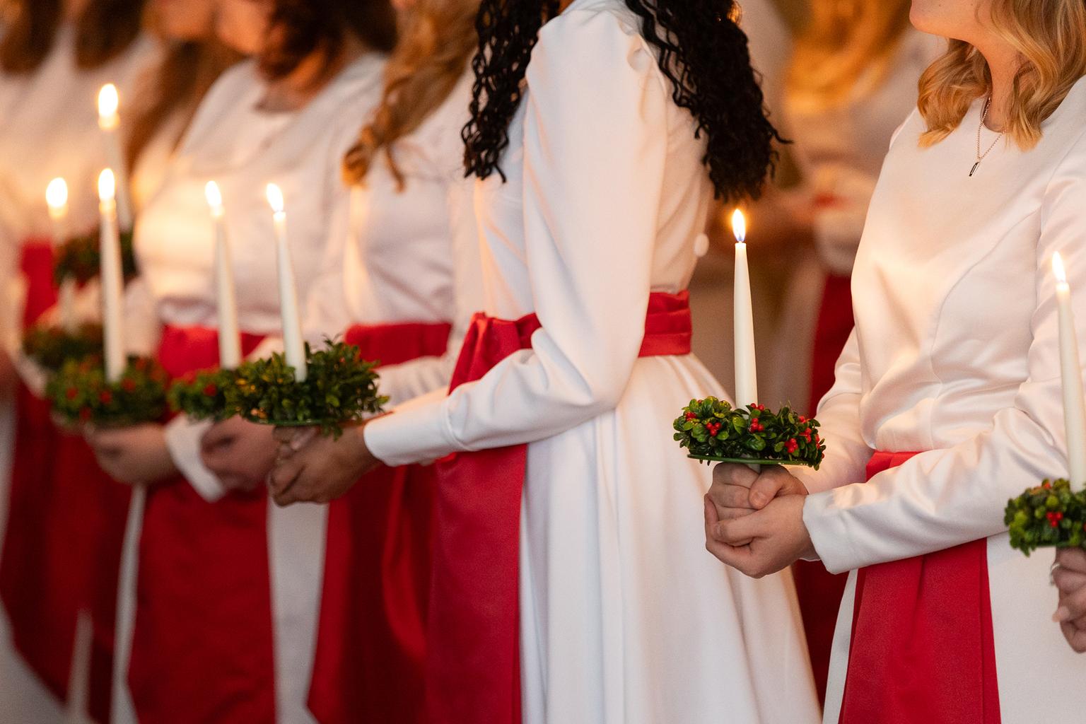 Lucia med tärnor på uppdrag med tända ljus. På bilden ser vi bara händerna som håller i ljusen, samt luciatågets klänningar, men inga ansikten. 