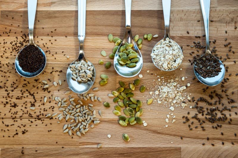 Fem skedar fyllda med olika frön och gryn, inklusive chiafrön, solrosfrön, pumpafrön och havre, arrangerade på en träyta.