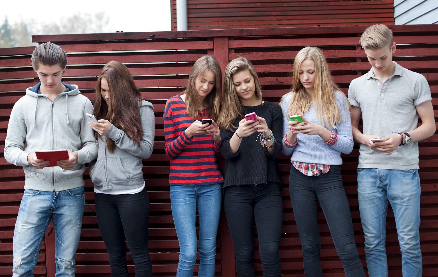 En grupp på sex tonåringar står lutade mot ett rött trästaket utomhus, alla fokuserade på sina mobiltelefoner och surfplattor. De är klädda i avslappnade kläder som jeans, hoodies och stickade tröjor. De ser koncentrerade ut och interagerar inte med varandra, vilket speglar dagens digitala vanor bland unga.