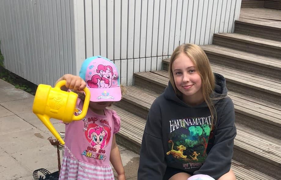 Therese Algar, nuori nainen, jolla on pitkät vaaleat hiukset ja tumma "Hakuna Matata" -huppari, istuu puuportailla ja hymyilee kameralle. Hänen vieressään seisoo pieni lapsi vaaleanpunaisessa mekossa ja värikkäässä lippalakissa, pitäen keltaista kastelukannua.