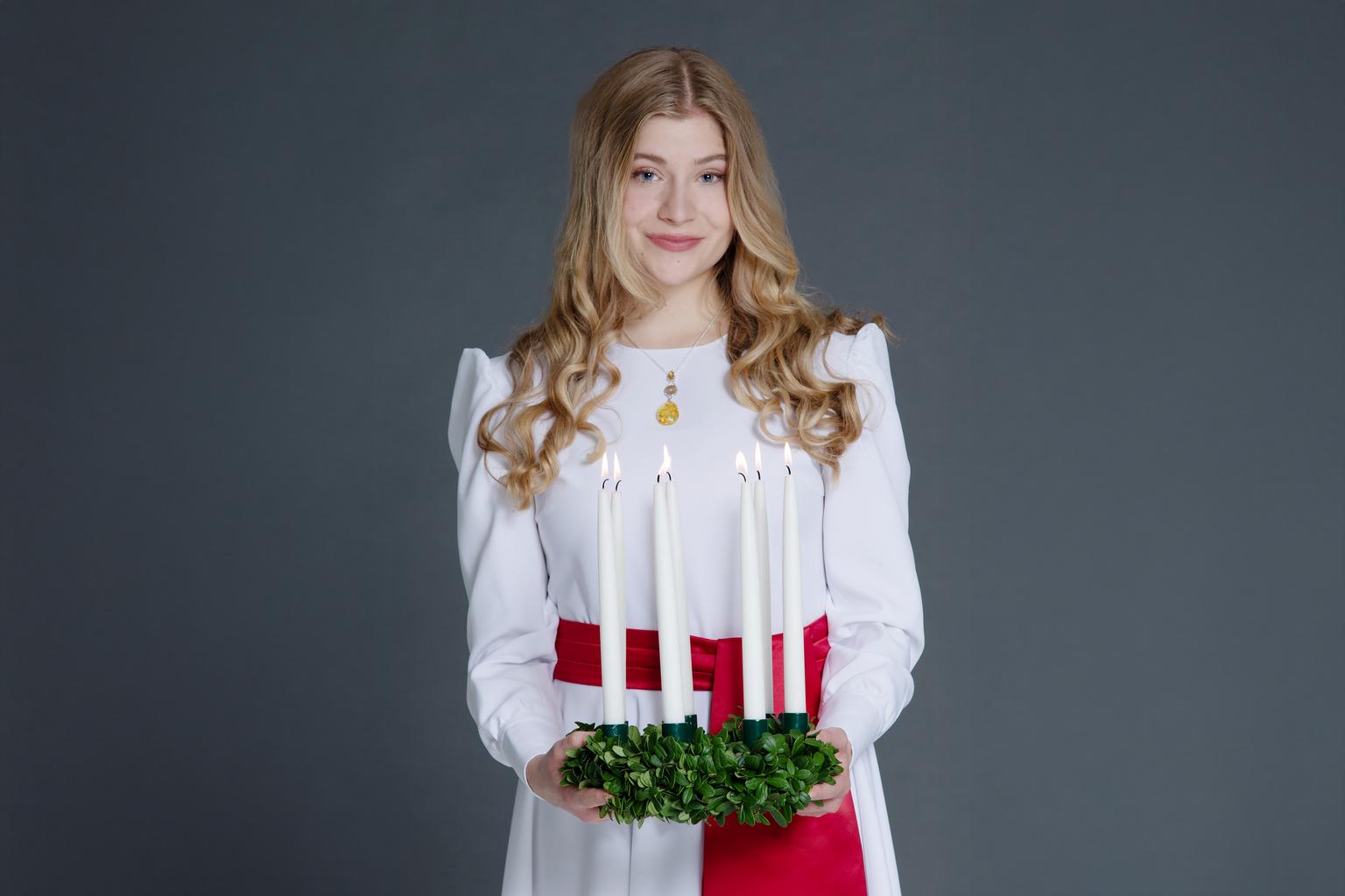 Finlands Lucia, klädd i vit skrud med rött band, håller leende fram en luciakrona med brinnande ljus i. 