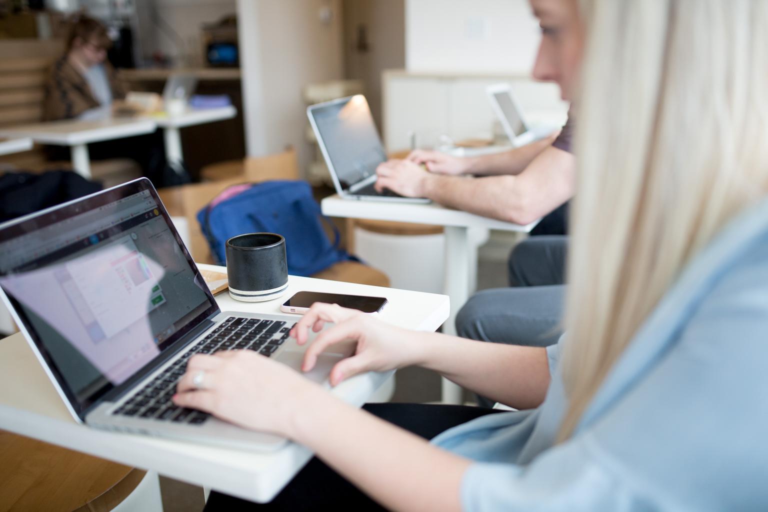 En ung kvinna arbetar på en laptop vid ett bord på ett café, omgiven av andra personer som också arbetar på sina datorer. Det är en ljus och modern miljö med en avslappnad atmosfär