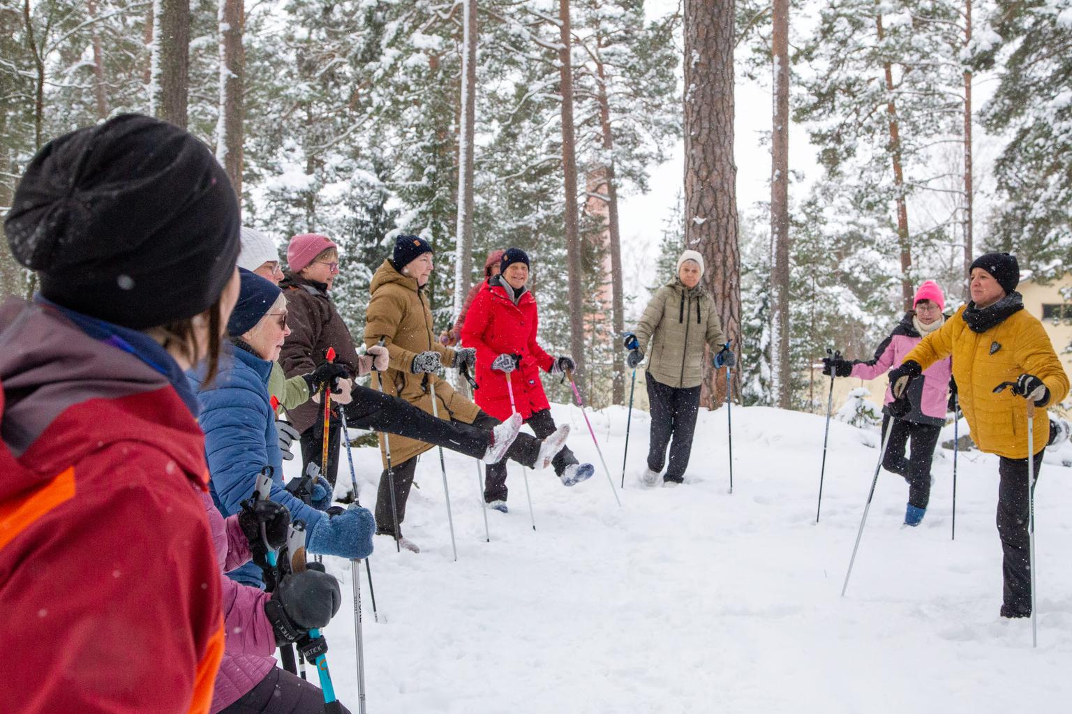 En grupp personer tränar med stavar i en vinterskog, omgivna av snö och träd.