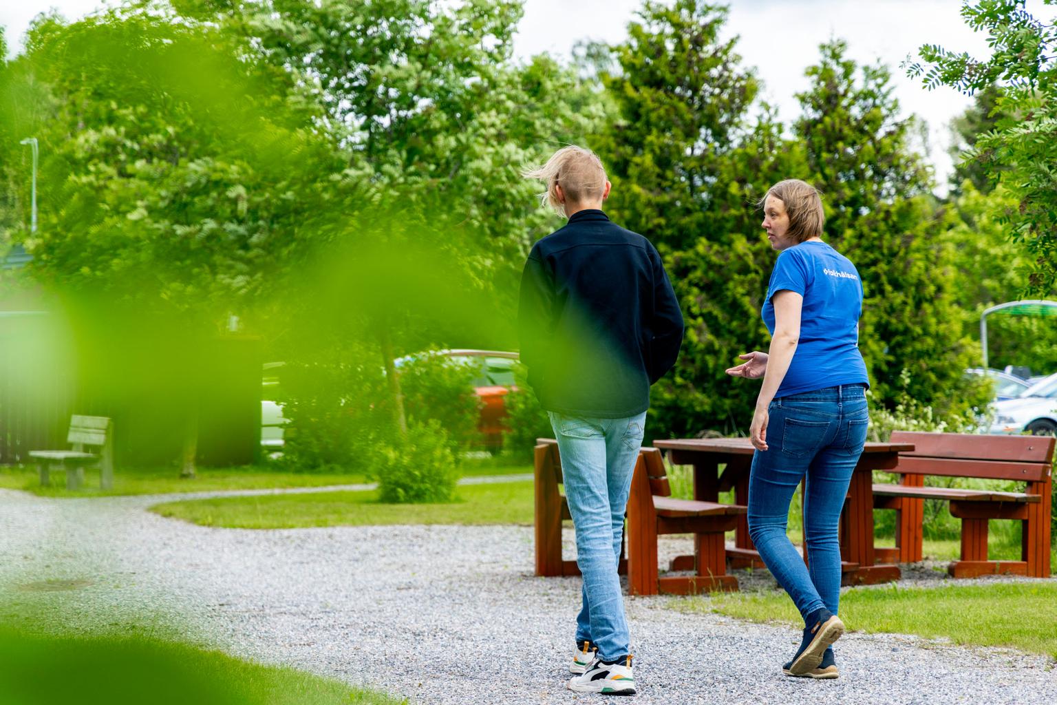 En ungdom och en vuxen promenerar längs en grusväg i en grönskande parkmiljö. Den vuxna, klädd i en blå Folkhälsan-tröja, gestikulerar medan de samtalar.