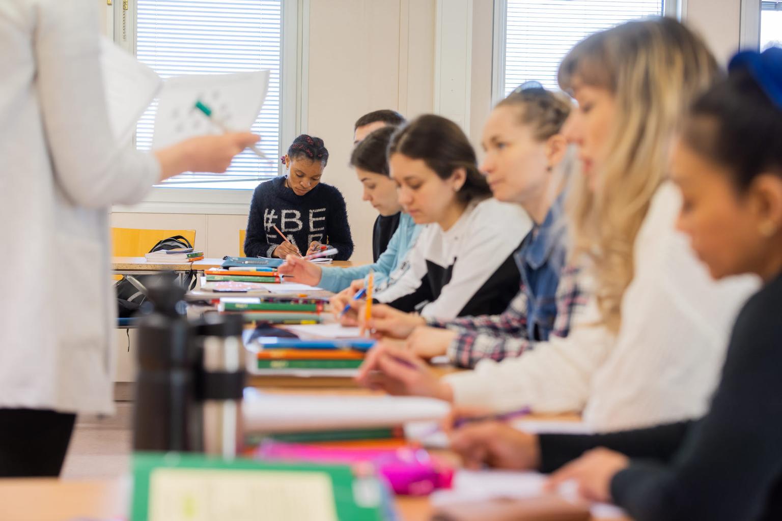 Opiskelijat luokkahuoneessa osallistuvat opetukseen, kirjoittavat muistiinpanoja ja kuuntelevat opettajaa. Värikkäitä kyniä ja muistivihkoja on pöydillä.