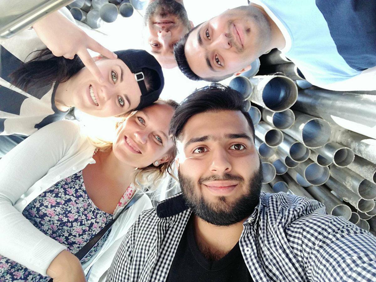 En grupp glada människor som tar en selfie, stående under ett konstverk av metallrör i ett utomhusområde.